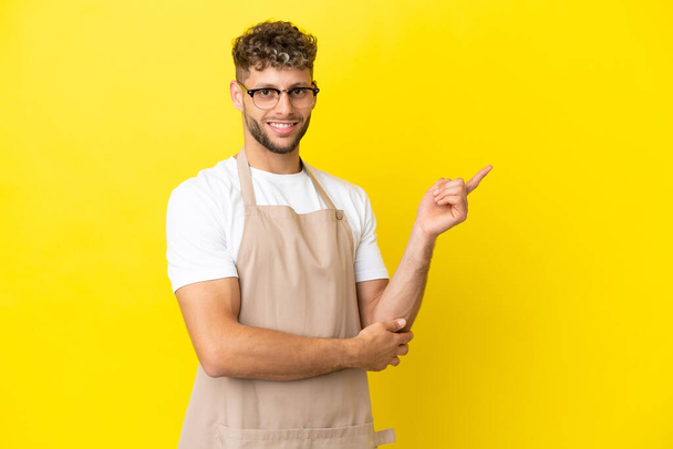 Restaurante camarero hombre rubio aislado sobre fondo amarillo señalando el dedo a un lado - Foto, imagen