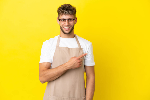 Εστιατόριο σερβιτόρος ξανθιά άνθρωπος απομονώνονται σε κίτρινο φόντο που δείχνει προς τα πλάγια για να παρουσιάσει ένα προϊόν - Φωτογραφία, εικόνα