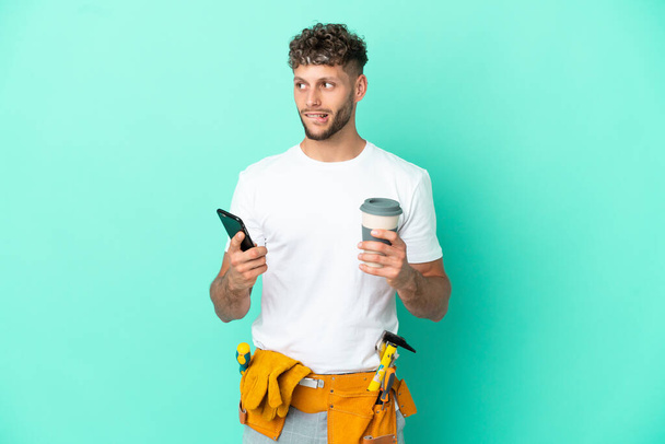 Молодой электрик блондин изолирован на зеленом фоне держа кофе, чтобы забрать и мобильный, думая что-то - Фото, изображение