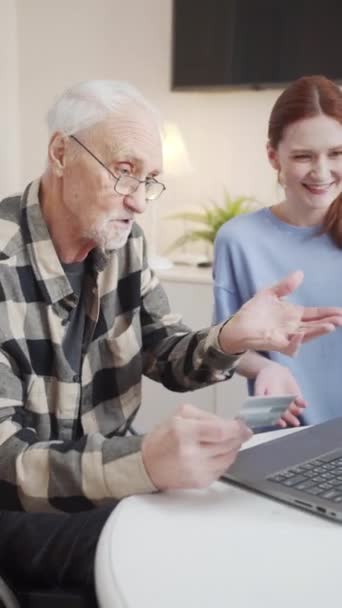 Een oude man in een rolstoel en een jonge vrouw zitten naast een laptop. Een gepensioneerde probeert te betalen voor aankopen op het internet met een bankkaart. Als hij slaagt, verheugt hij zich, en het meisje kalmeert - Video