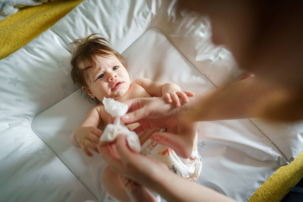 Un pequeño bebé caucásico acostado en la cama desnudo con manos de madre mujer desconocida usando toalla de servilleta húmeda para limpiar su piel mientras crece el cuidado de la higiene infantil y el concepto de paternidad - Foto, Imagen