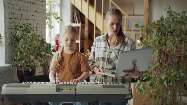 Dziecko naciska klawisze pianina elektronicznego. Jej matka stoi w pobliżu i trzyma laptopa buka, na którym notatki są otwarte.. Wysokiej jakości materiał 4k - Materiał filmowy, wideo