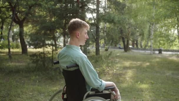 Een jonge gehandicapte man in een blauw shirt zit in de rolstoel, kijkt weg en geniet van het weer. Hoge kwaliteit 4k beeldmateriaal - Video