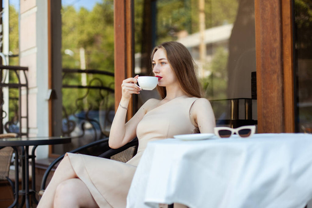 若い女性がコーヒーを飲みながら屋外カフェのテーブル席に座る - 写真・画像