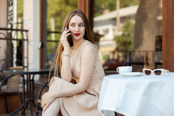 Jovem mulher elegante falando ao telefone enquanto se senta em uma mesa em um café - Foto, Imagem