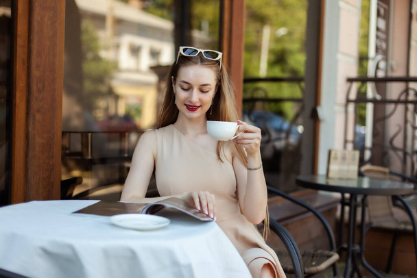 Young fashion woman reading a magazine while sitting at table in outdoor cafe and drinking coffee - Foto, afbeelding