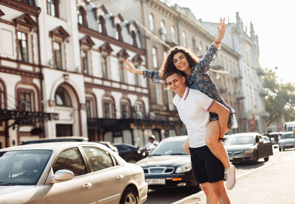 Couple in love. Man carrying girl on his back in the street. Smiling man with beautiful young woman, ride piggyback, having fun together. Relationship concept. - Фото, изображение