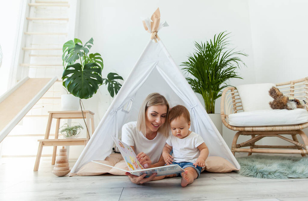 Happy woman mom and her little daughter read book in wigwam at home - Фото, зображення