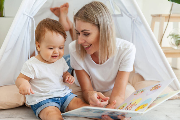 Happy woman mom and her little daughter read book in wigwam at home - Foto, imagen