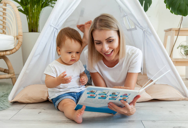 Happy woman mom and her little daughter read book in wigwam at home - Zdjęcie, obraz