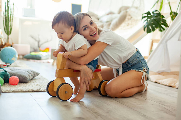 Mom and little daughter spend time in the playroom. Little girl on a wooden scooter. Maternal care, love concept - Photo, Image