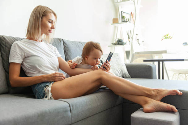Милая молодая мама со своей маленькой дочерью пользуется смартфоном, сидя дома на диване. - Фото, изображение