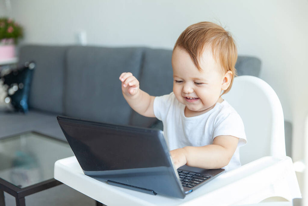 Kleines lustiges Baby benutzt Laptop, während es zu Hause am Kindertisch sitzt - Foto, Bild