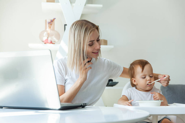 Evden çalışıyorum. Genç anne bir dizüstü bilgisayarla masada oturup kızını beslerken telefonla konuşuyor. Annelik, annelik, çocukluk ve bakım kavramı. - Fotoğraf, Görsel