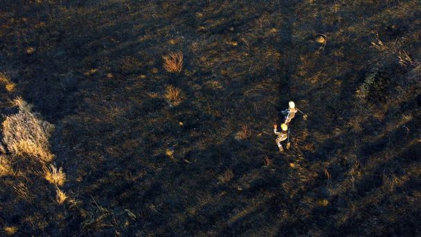 Два пожарных ходят по черной выжженной земле после пожара сухая трава в поле. - Фото, изображение