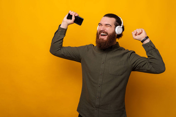Чудовий бородатий чоловік танцює, слухаючи музику в навушниках. Студійний знімок на жовтому тлі
. - Фото, зображення