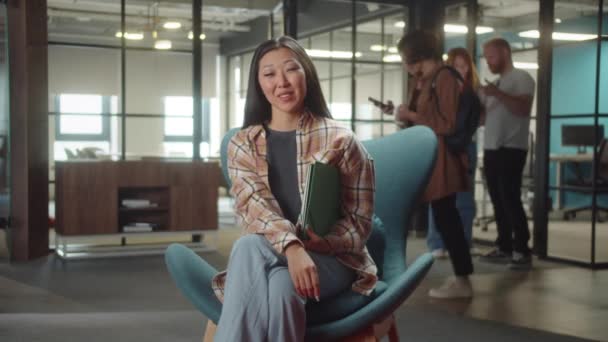 Genç Asyalı bir bayan üniversitede sandalyede oturuyor, gülümsüyor ve kameraya bakıyor. Yüksek kaliteli FullHD görüntüler - Video, Çekim