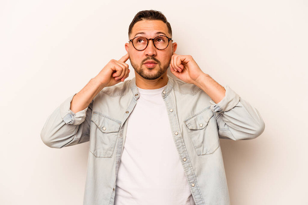 Young hispanic man isolated on white background covering ears with fingers, stressed and desperate by a loudly ambient. - Foto, imagen