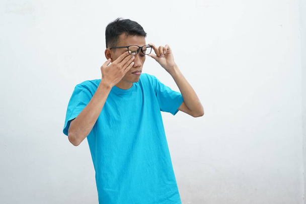 Jeune asiatique portant un t-shirt bleu malsain avec des lunettes, des yeux frottés, souffrant de vision floue, syndrome des yeux secs, vertiges, isolé sur fond blanc. Concept de problème de santé - Photo, image