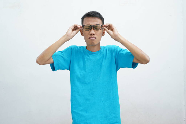 Mladý Asiat nosí nezdravé modré tričko s brýlemi, tření očí, trpí rozmazaným viděním, syndromem suchého oka, závratí, izolované na bílém pozadí. Koncept zdravotního problému - Fotografie, Obrázek