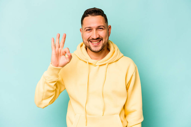 Jonge Spaanse man geïsoleerd op blauwe achtergrond vrolijk en vol vertrouwen tonen ok gebaar. - Foto, afbeelding