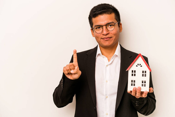Young business hispanic man holding a little house isolated on white background showing number one with finger. - Fotografie, Obrázek