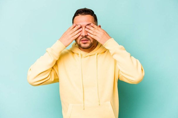 Young hispanic man isolated on blue background afraid covering eyes with hands. - 写真・画像
