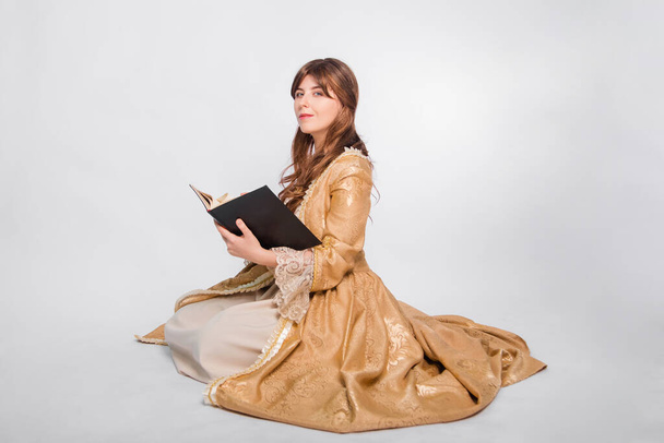 Full length portret van een jonge vrouw in een gezwollen gouden jurk in de rococo tijdperk, poseren terwijl ze zit met een boek in haar handen, geïsoleerd op een witte achtergrond. Het meisje leest een boek.. - Foto, afbeelding