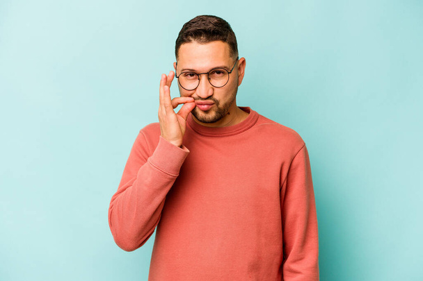 Young hispanic man isolated on blue background with fingers on lips keeping a secret. - 写真・画像