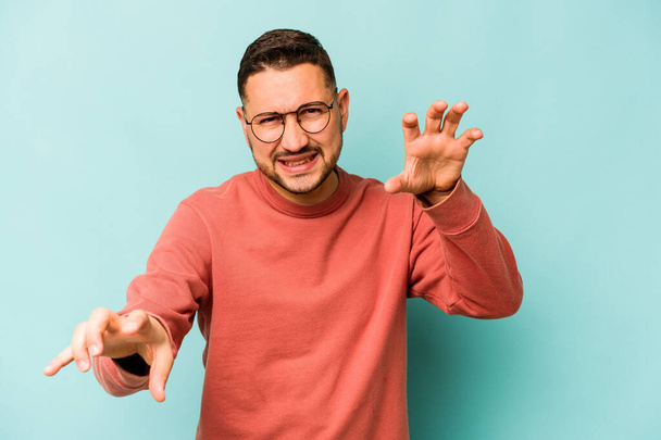 Young hispanic man isolated on blue background showing claws imitating a cat, aggressive gesture. - Photo, Image