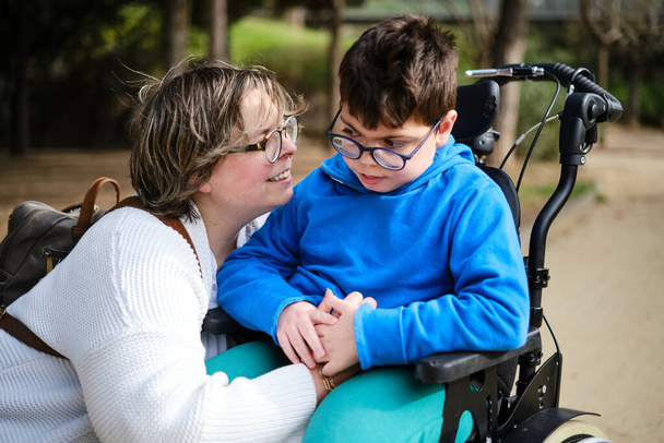 Disabled boy in a wheelchair enjoying a walk outdoors with his mother. - Zdjęcie, obraz