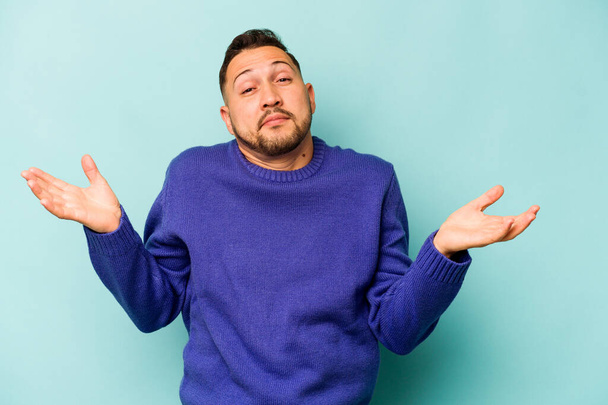 Young hispanic man isolated on blue background doubting and shrugging shoulders in questioning gesture. - Photo, Image
