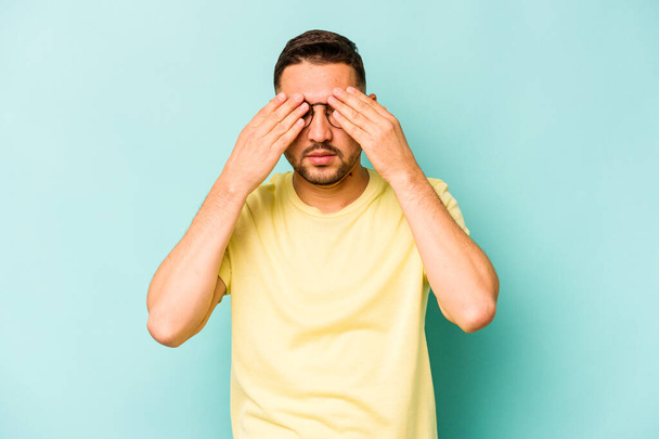 Jonge Spaanse man geïsoleerd op blauwe achtergrond bang voor het bedekken van ogen met handen. - Foto, afbeelding