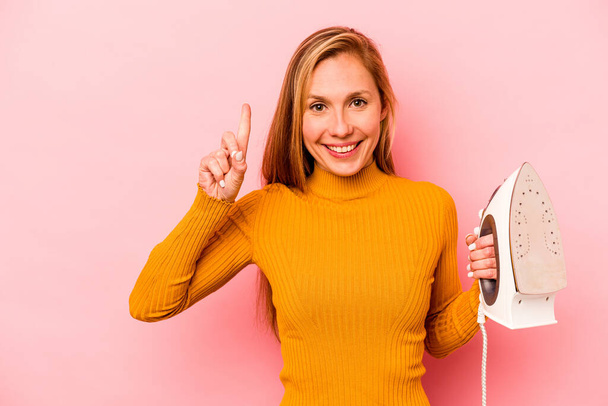 Young caucasian woman holding iron isolated on pink background showing number one with finger. - Photo, Image