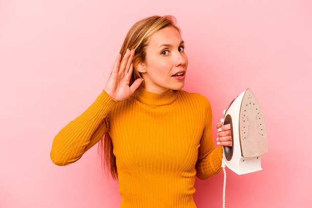 Молодая белая женщина, держащая железо на розовом фоне, пытается слушать сплетни. - Фото, изображение