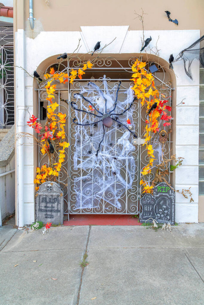 Schmiedeeisernes Tor mit Holloween-Dekorationen in San Francisco, Kalifornien. Es gibt eine große Plüschspinne, falsche Reben, Krähen und Fledermäuse an der Tür mit Betonplatte Eingang. - Foto, Bild