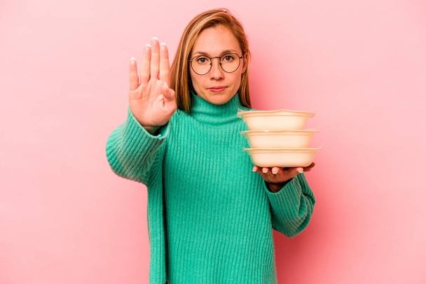 Jonge blanke vrouw houden tupperware geïsoleerd op roze achtergrond staan met uitgestrekte hand tonen stop teken, voorkomen dat u. - Foto, afbeelding
