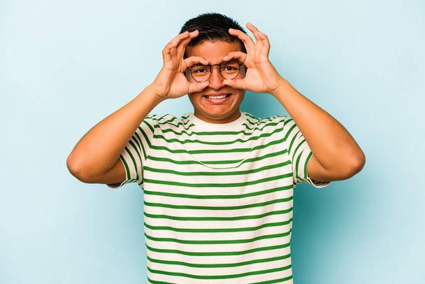 Jonge Spaanse man geïsoleerd op blauwe achtergrond toont goed teken over de ogen - Foto, afbeelding