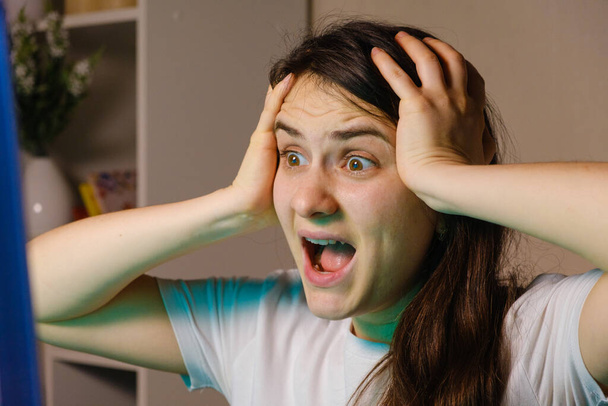 A woman screams and grabs her head with her hands while looking at a TV or computer screen. - Photo, Image