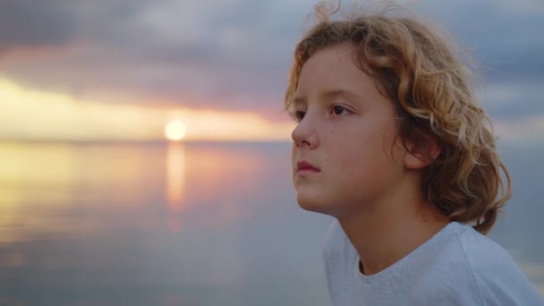 Çekici çocuk güzel bir günbatımının arka planında kameraya bakar. Yakın plan. - Video, Çekim
