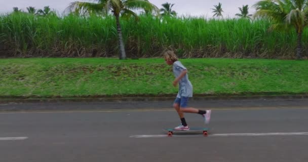 Un adolescent monte un longboard le long d'une belle route avec des palmiers verts - Séquence, vidéo