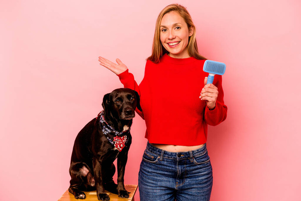 Young caucasian woman combing the dog isolated on pink background showing a copy space on a palm and holding another hand on waist. - Zdjęcie, obraz