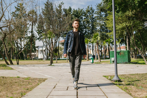 Front view of young man in jeans clothes slowly walking through alley of trees in nice day at park. Spending time alone in nature. Peaceful atmosphere. - 写真・画像