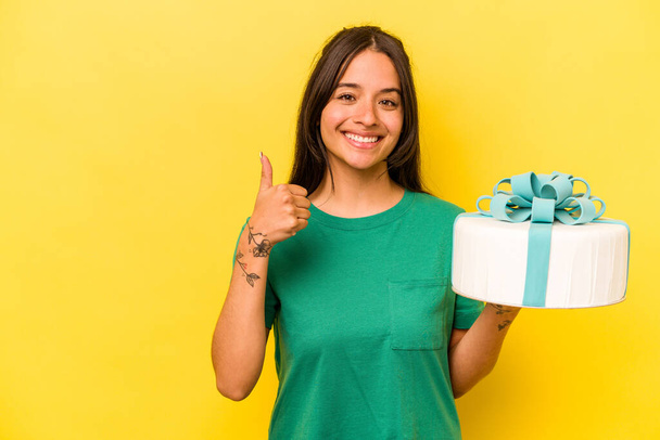 Young hispanic woman holding birthday cake isolated on yellow background smiling and raising thumb up - Fotó, kép
