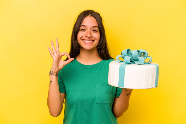 Молода іспаномовна жінка тримає торт на день народження ізольовано на жовтому тлі веселий і впевнений, показуючи добре жест
. - Фото, зображення