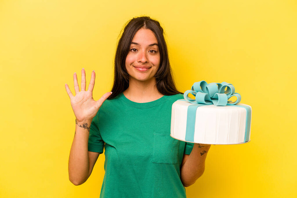 Jonge Spaanse vrouw met verjaardagstaart geïsoleerd op gele achtergrond glimlachend vrolijk tonen nummer vijf met vingers. - Foto, afbeelding