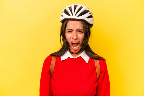 Joven estudiante hispana usando un casco de bicicleta aislado sobre fondo amarillo gritando muy enojado y agresivo. - Foto, Imagen