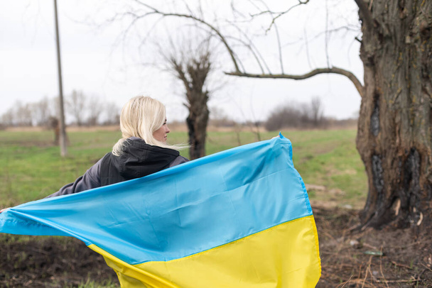 Mujer sosteniendo una bandera amarilla y azul de Ucrania sobre un fondo de árbol quemado - Foto, imagen