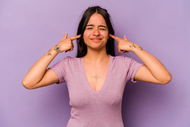 若いヒスパニック系の女性は、指で耳を覆う紫色の背景に隔離され、大声で周囲に強調され、絶望的な. - 写真・画像