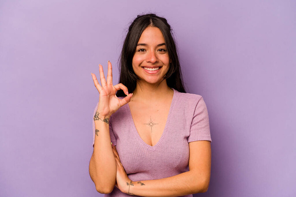 Молода іспаномовна жінка ізольована на фіолетовому фоні виграє око і тримає добре жест рукою
. - Фото, зображення